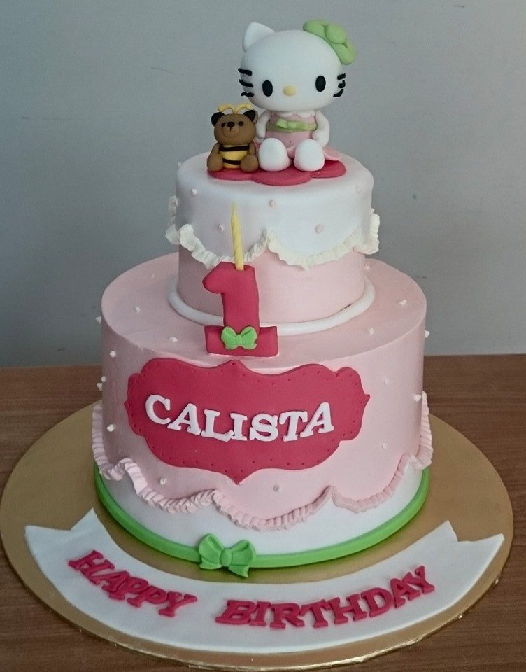 A pink two-tiered cake with Hello Kitty figure for baby girl’s first birthday. Made by: My Fat Lady Cakes and Bakes. Source