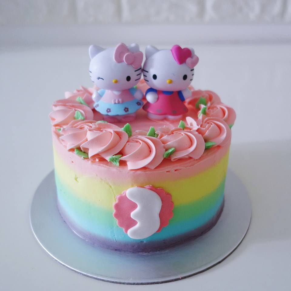 A tall round cake with rainbow buttercream frosting and Hello Kitty figures. Made by: River Ash Bakery. Source