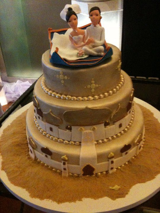A white and gold wedding cake with figurines of the newlyweds flying on a magic carpet is perfect for an Aladdin themed wedding. Made by: Bonheur Patisserie.Source
