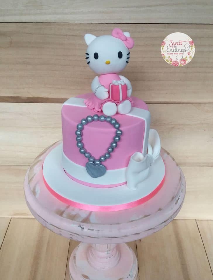 A simple, round cake with fondant icing, decorated with with a simple bow on the side, grey fondant shaped like a necklace, and a cute-looking Hello Kitty figure on top. Made by: Sweet Endings.Source